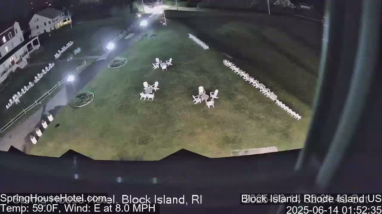 A nighttime, overhead view of a grassy area with several small groups of white chairs and tables, a walkway, and a building in the background under a clear sky.