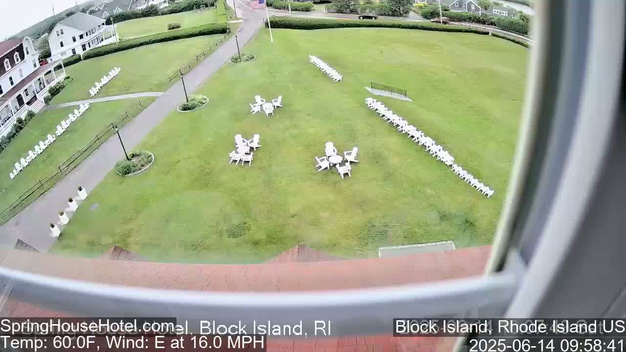 A grassy lawn with several rows of white chairs and small groupings of tables and chairs, viewed from an elevated position on a partly cloudy day with an east wind.
