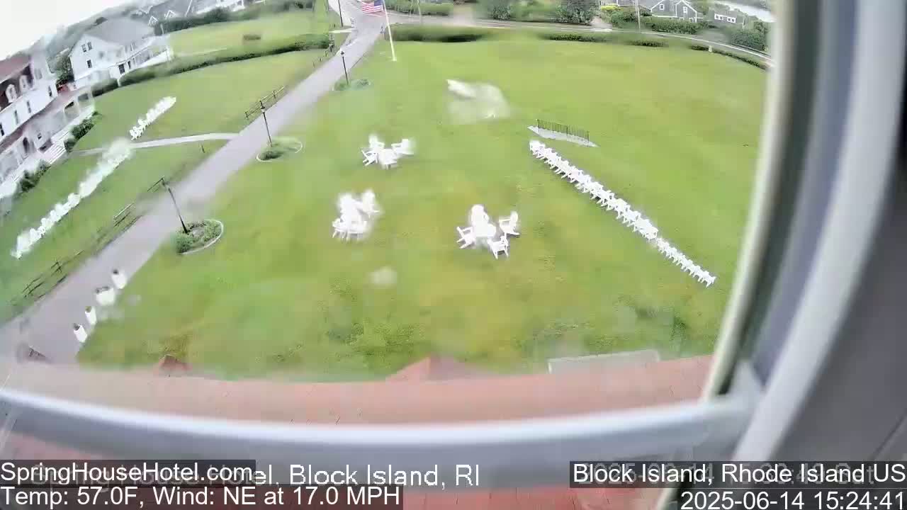 A grassy lawn with several rows of white chairs and small groupings of tables and chairs, viewed from an elevated position on a partly cloudy day with an east wind.