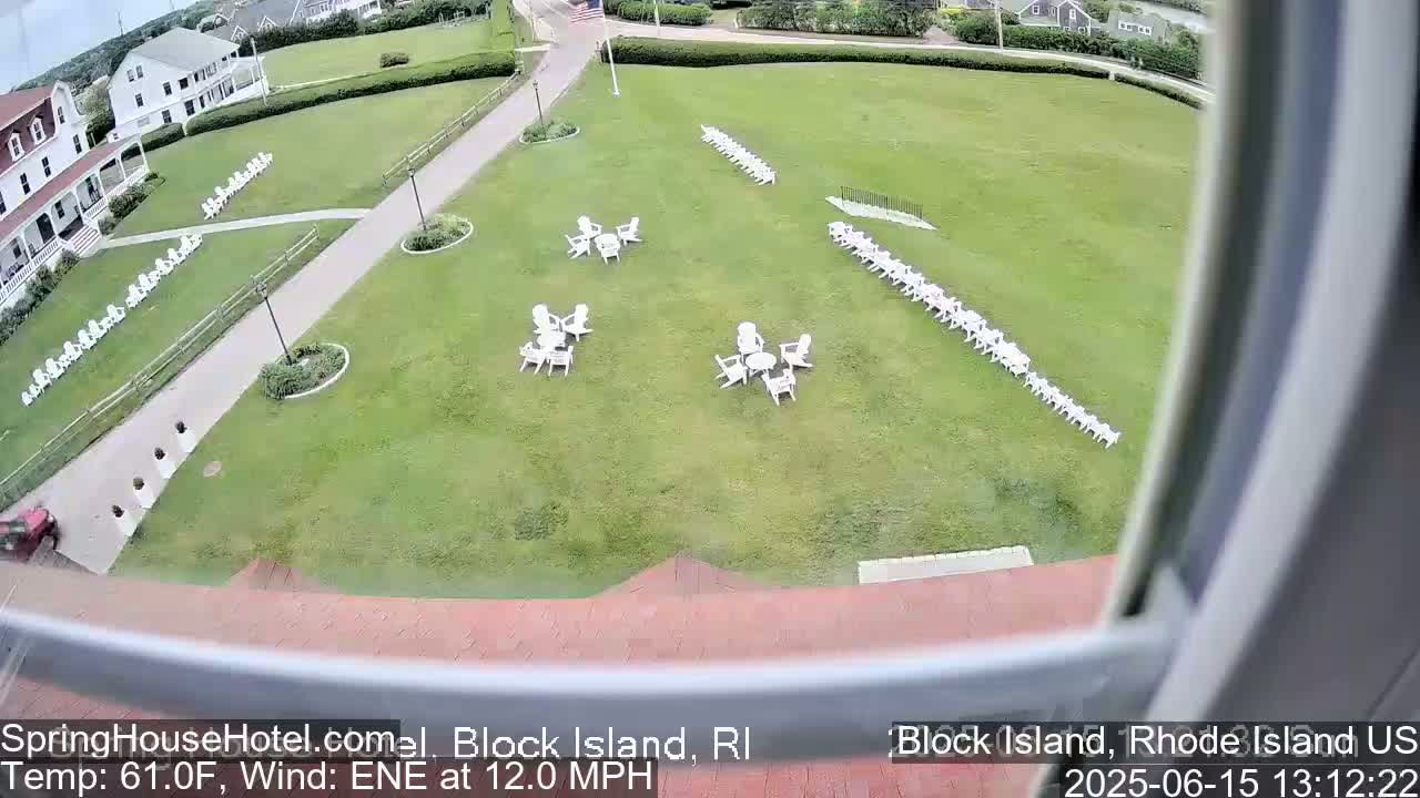A grassy lawn with several sets of white chairs and small tables is seen from an elevated vantage point, with a building and a paved path visible in the background, in windy conditions.