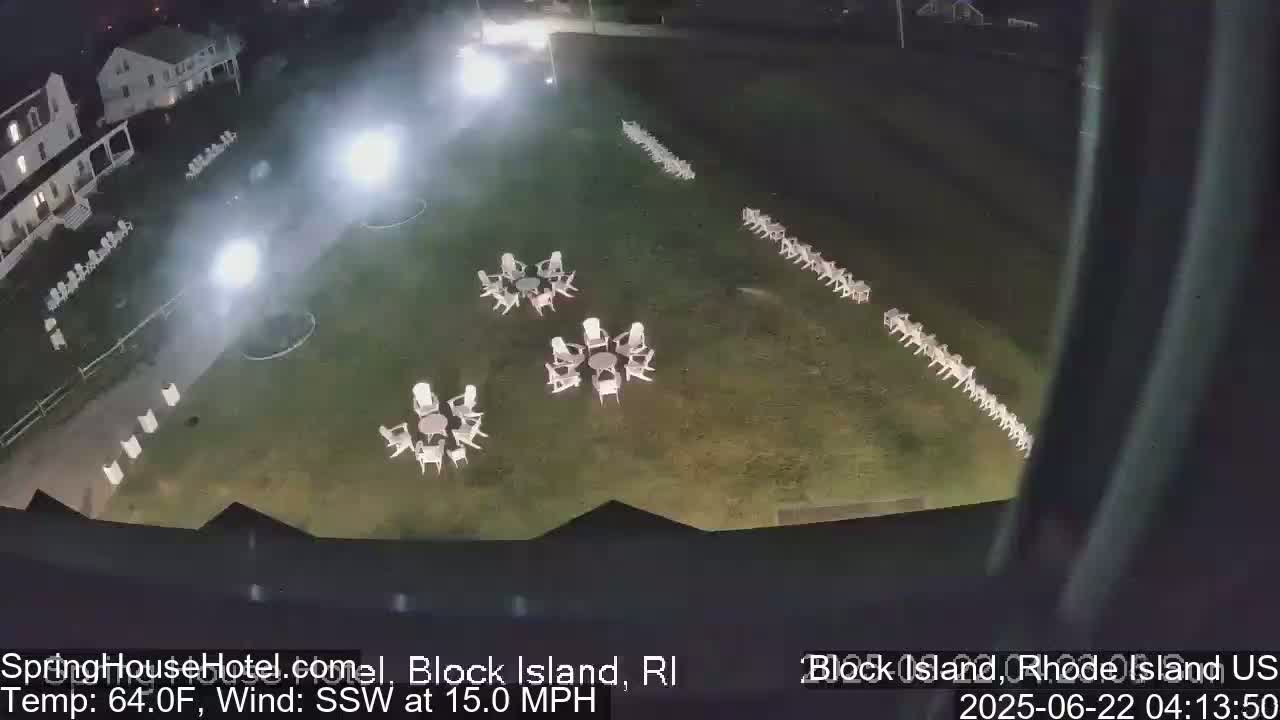 A nighttime, high-angle view shows a grassy area with numerous white chairs arranged in groups and a long line, illuminated by bright lights under a dark sky with a 15 mph south-southwesterly wind and a temperature of 64°F.