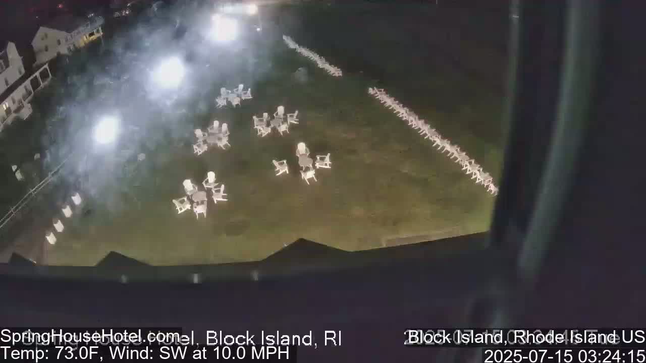 An aerial nighttime view shows a grassy area with numerous empty white chairs arranged in clusters and long rows, lit by artificial lights under a partly cloudy sky with a southwest wind.