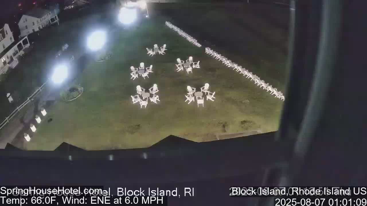 A nighttime aerial view shows a grassy area with numerous white chairs and tables arranged in clusters and a long row, illuminated by bright lights, with a building visible in the background under a clear sky.