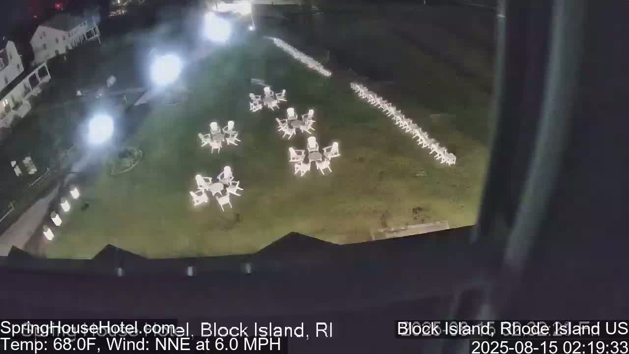 A nighttime, high-angle view shows a grassy area with numerous white tables and chairs arranged in groups and a long line, under a partly cloudy sky with a gentle north-northeast wind.