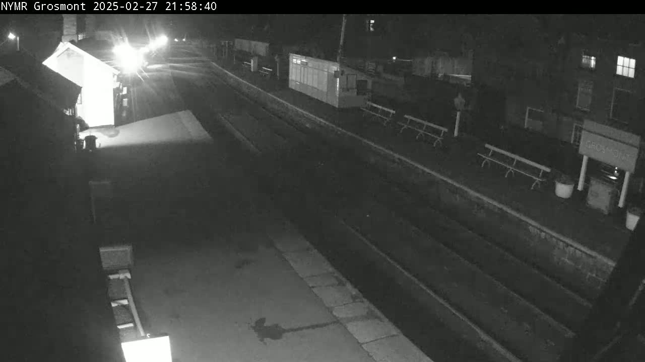 A nighttime, black-and-white view of a train station platform with several benches and small buildings under dark skies.