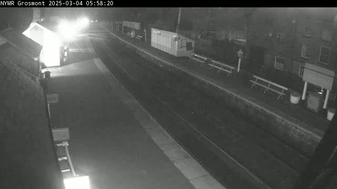 A nighttime view of a mostly empty train station platform with a few benches and buildings visible under dark conditions.