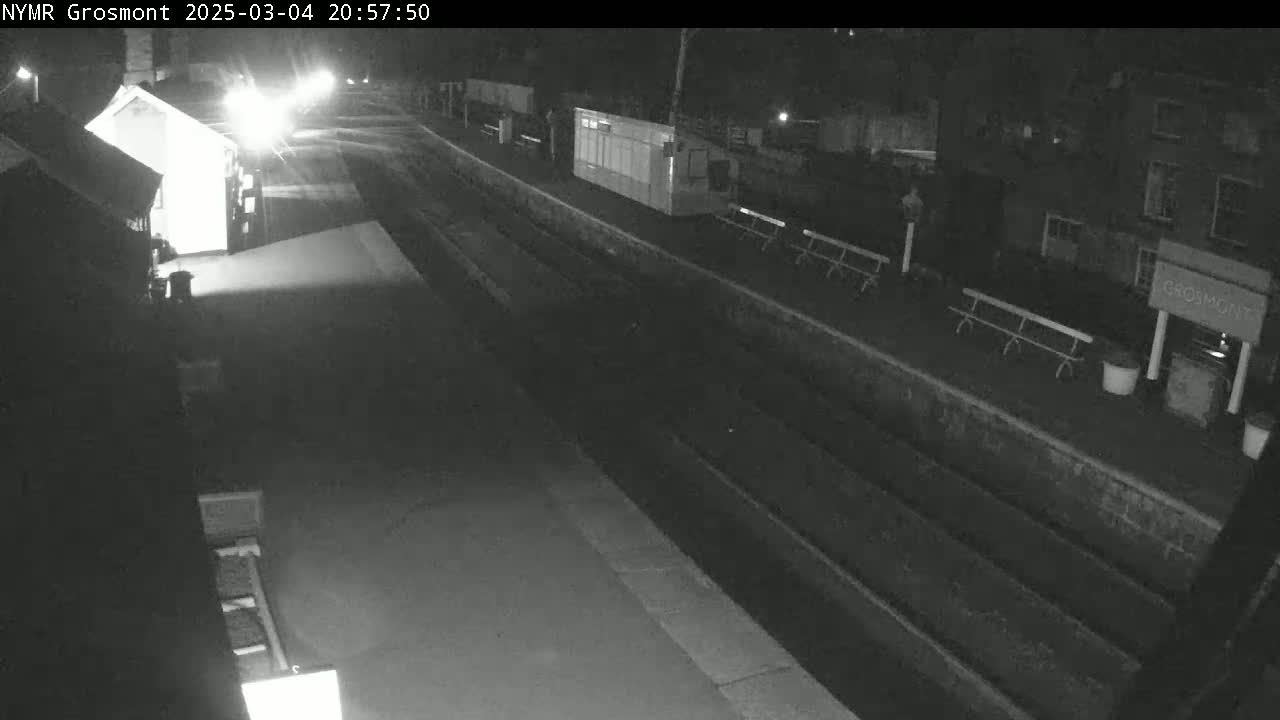 A nighttime, low-light view of a mostly empty train station platform with several benches and small buildings.