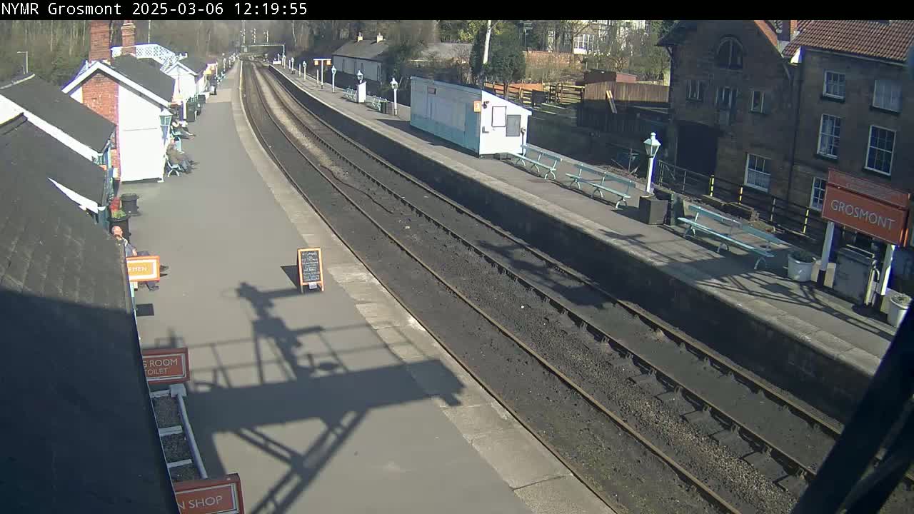 A sunny day at a train station with multiple tracks, several small buildings lining the platform, and benches along the edge.