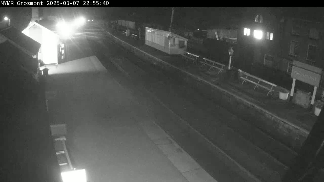 A nighttime, black-and-white view of a mostly empty train station platform with a building and benches visible.