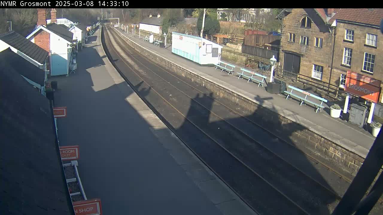 A sunny view of a train station platform with two parallel train tracks, several small buildings, and benches.