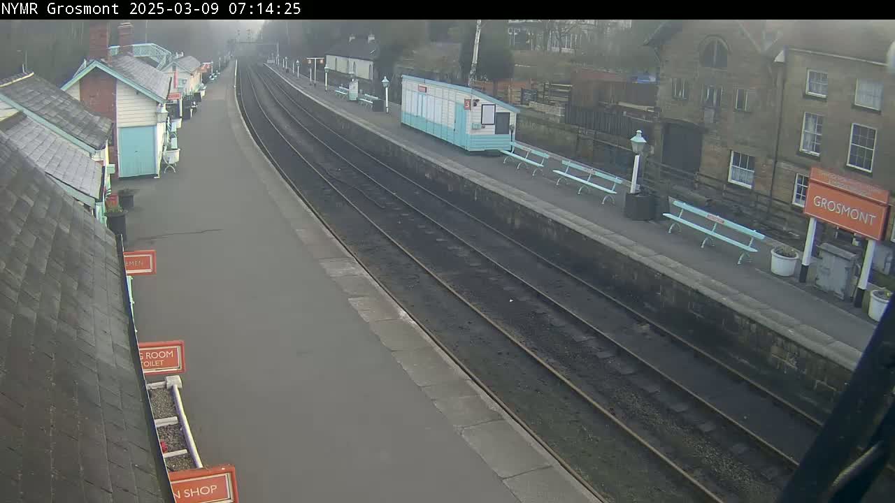 A mostly empty train platform on a foggy day with several small buildings and benches lining the platform next to the train tracks.