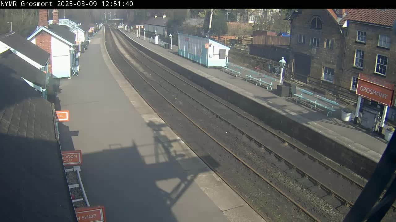 A sunny day at a train station with two parallel train tracks, several small buildings, and benches on the platform.