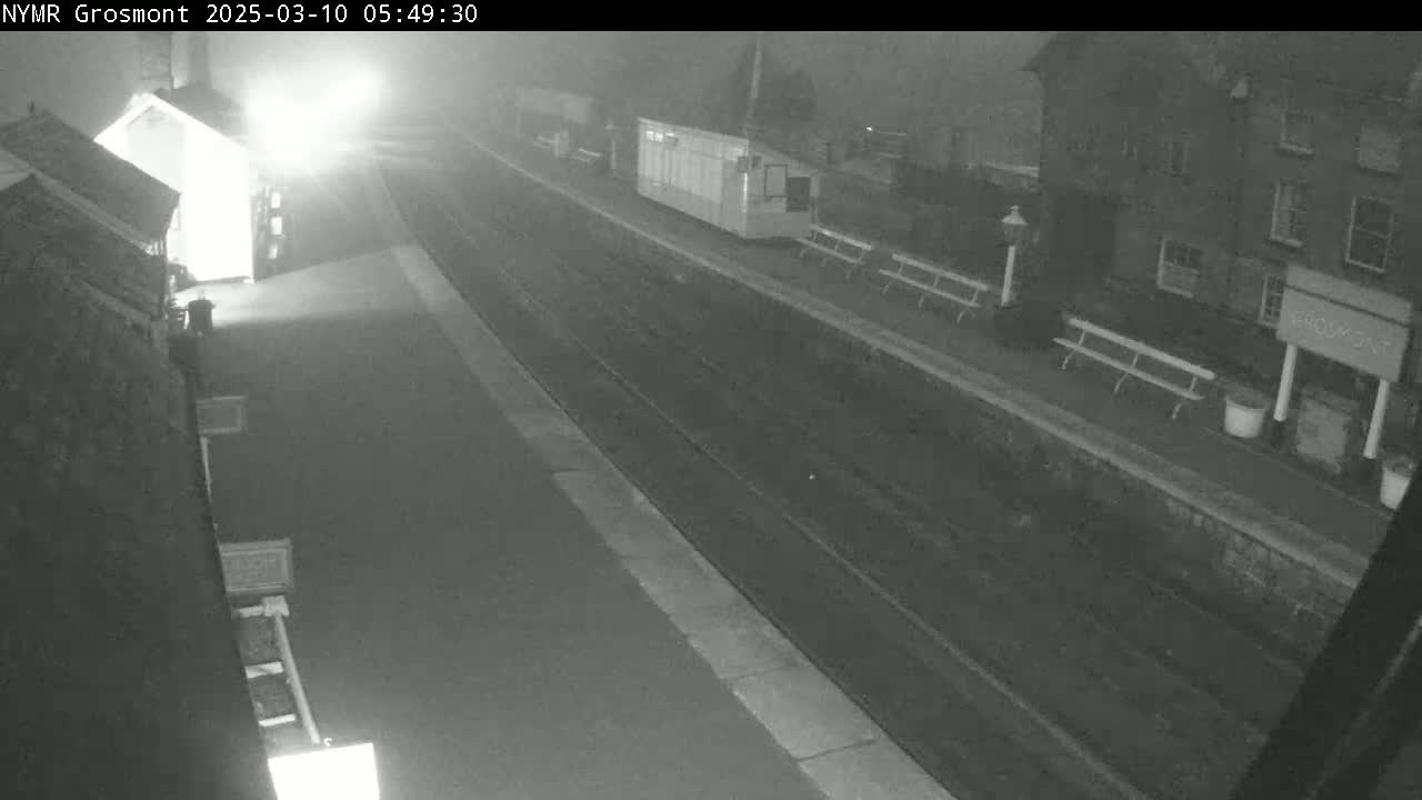 A nighttime, foggy view of a train platform with buildings and benches visible.