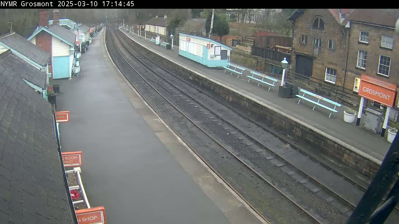 A wet train station platform with two tracks, several small buildings, and light-blue benches is seen on an overcast day.