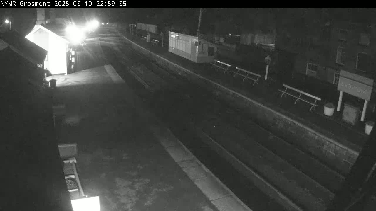 A nighttime, low-light view of a train station platform with benches and buildings, showing a mostly empty platform and a road in the background.