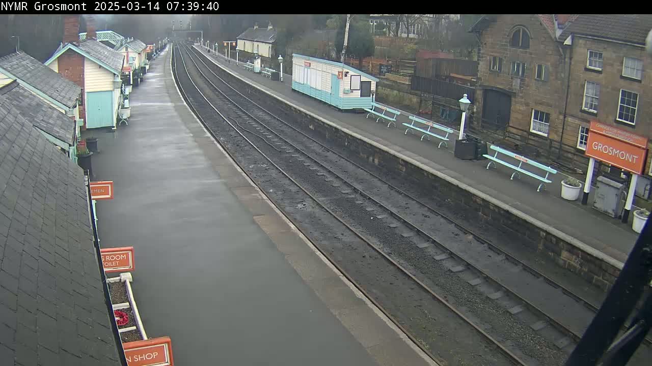 A wet train station platform with two sets of train tracks, several small buildings, and light-teal benches.