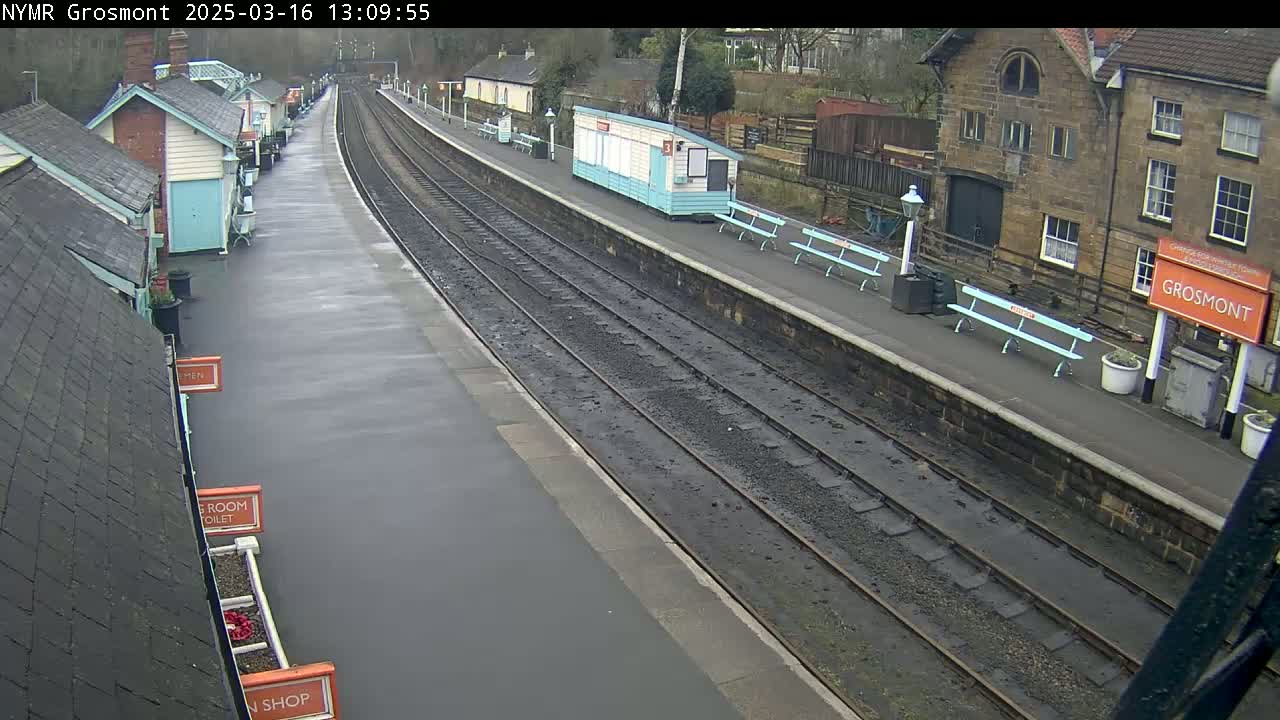 A wet train station platform with multiple tracks, several small buildings, and benches.
