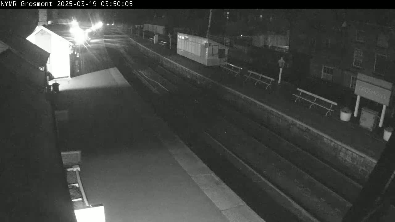 A nighttime view of a train station platform with train tracks, benches, and small buildings under dark conditions.