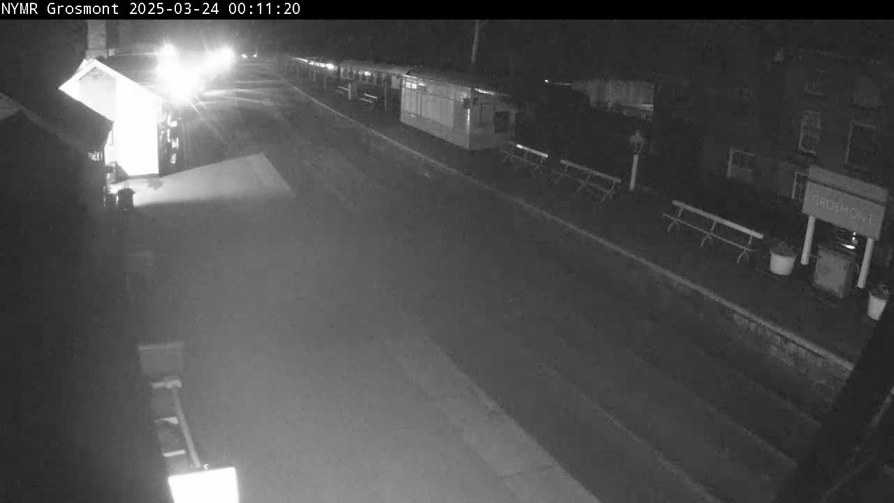 A nighttime, black and white view of a deserted road and train station platform with several benches and buildings.