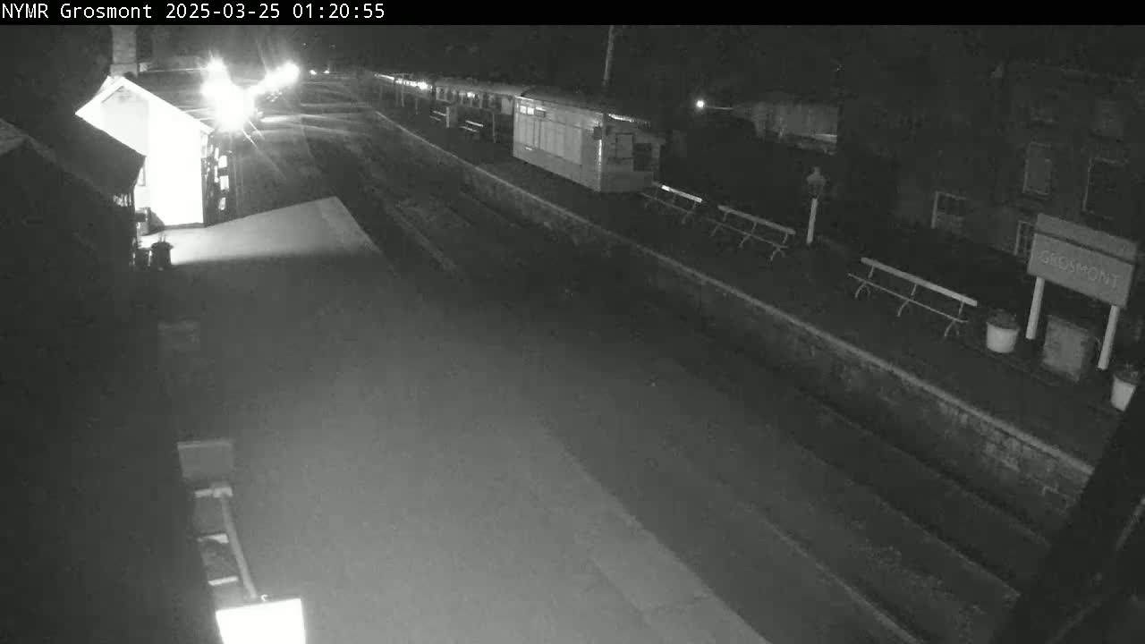 A nighttime, low-light view of a mostly empty train station platform with a few benches and buildings visible.