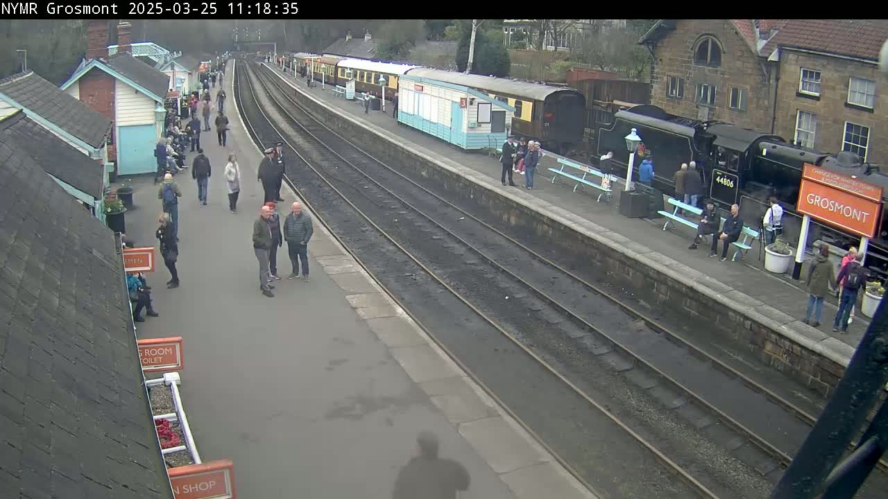 A train station with multiple train cars and many people standing around on a mostly sunny day.