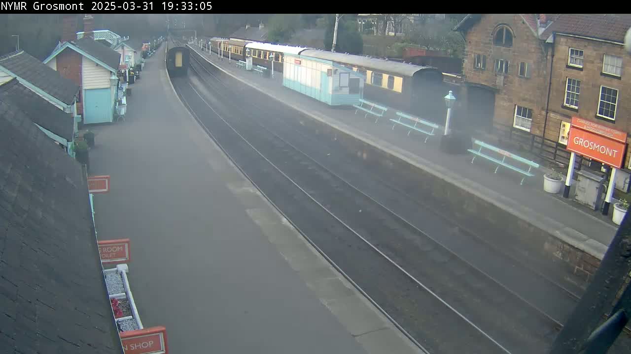 A misty train station with a long passenger train at the platform and several small buildings along the side of the tracks.