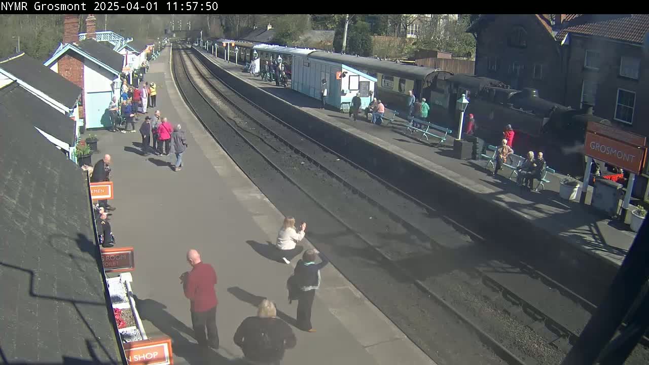 A sunny day at a train station shows a steam train at the platform with many people standing and sitting around, some taking photos.