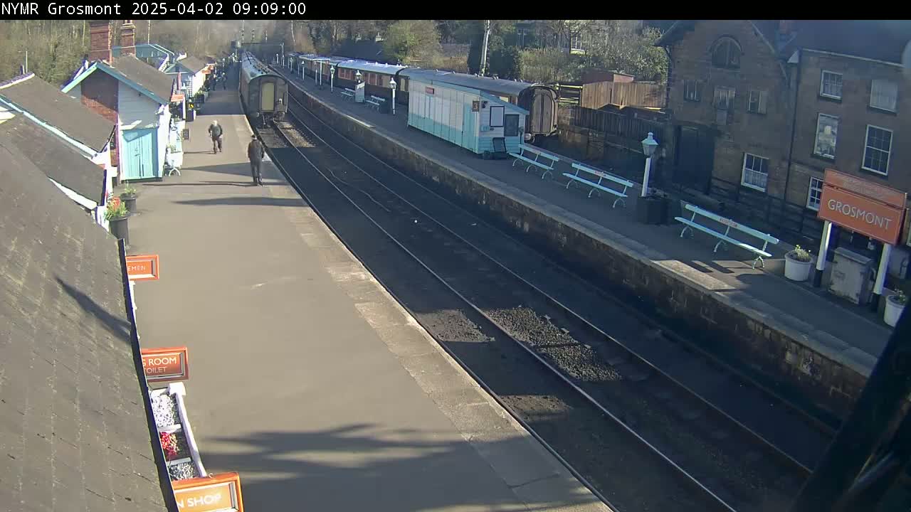 A sunny day at a train station shows several railway carriages and a few people walking on the platform, with small buildings nearby.