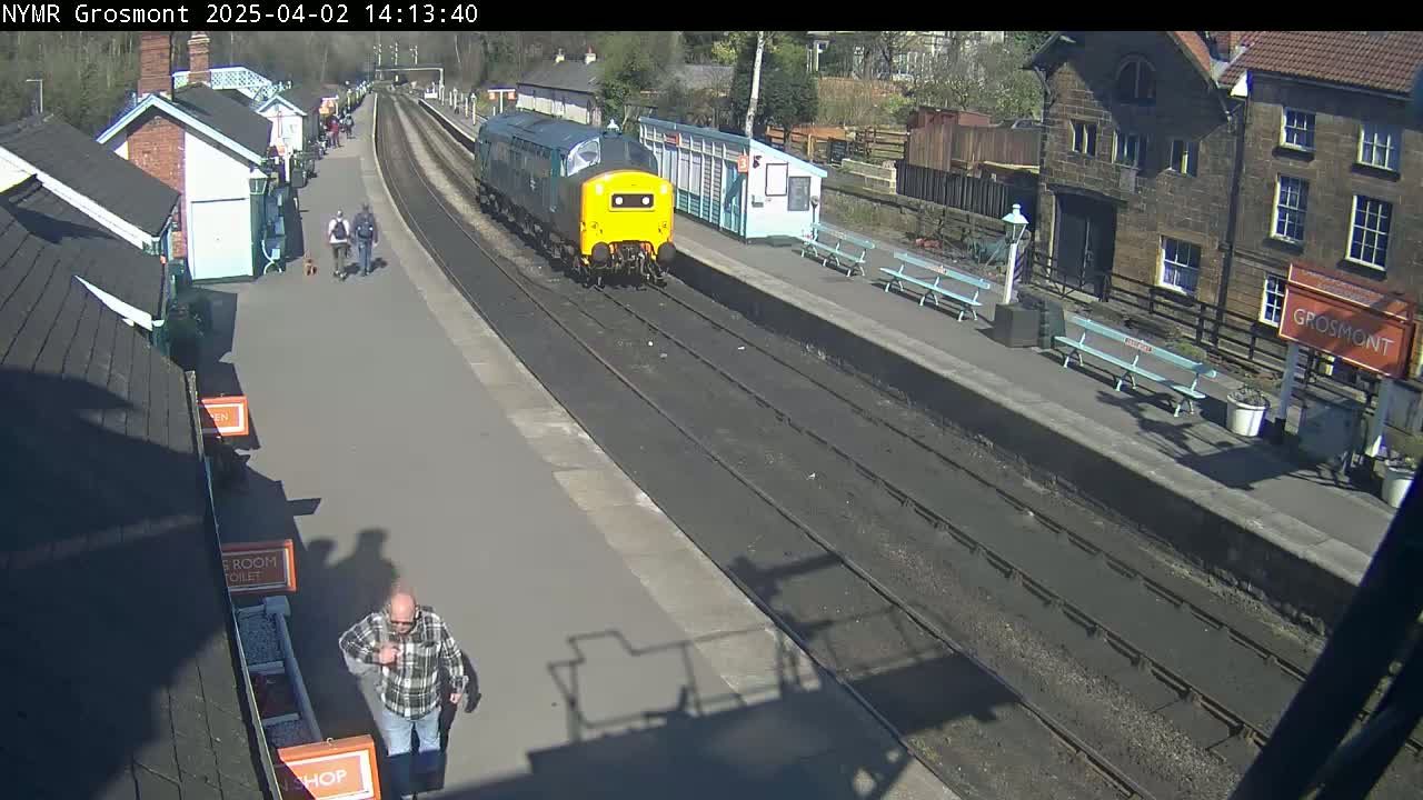 A yellow and blue diesel locomotive is arriving at a train station on a sunny day, with a few people walking along the platform.
