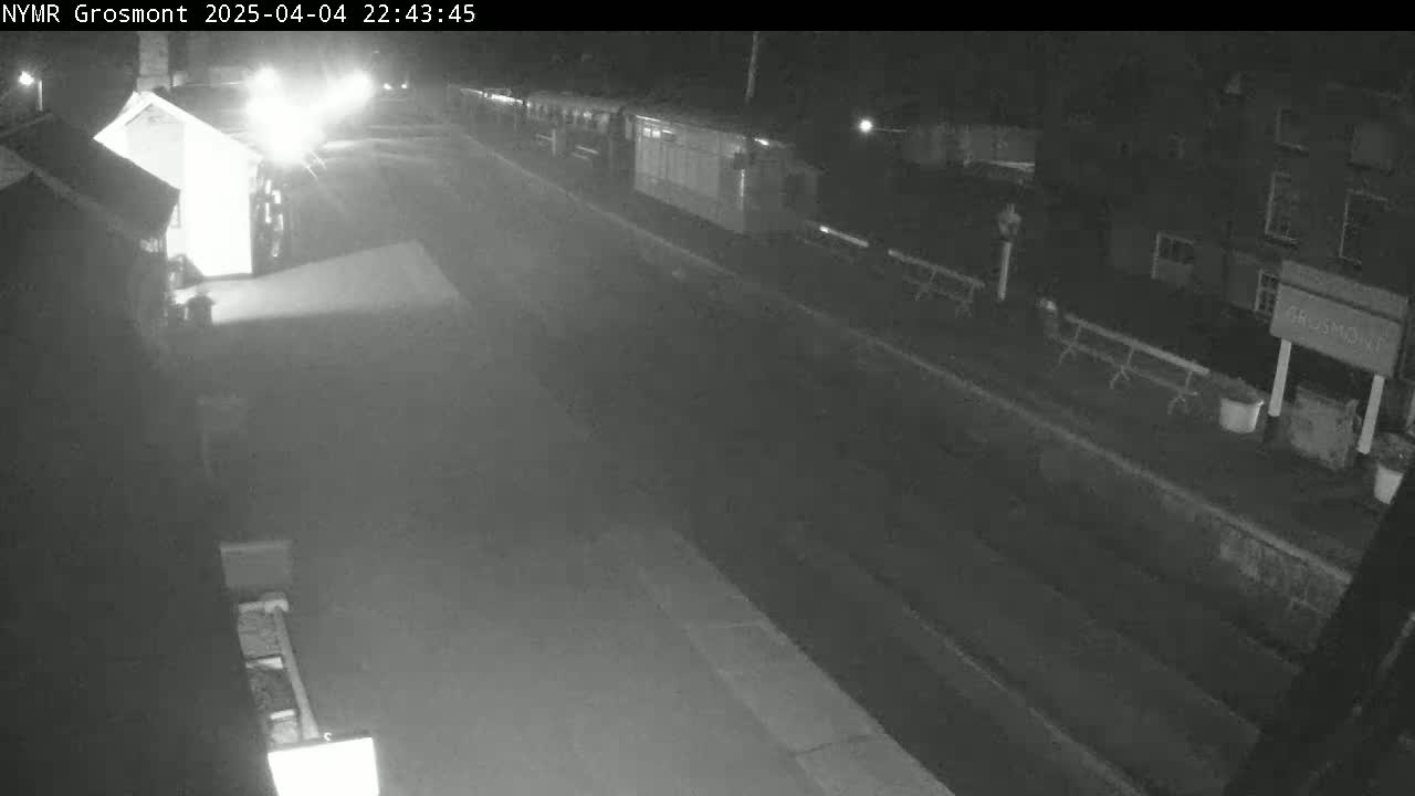 A long, dark roadway at night shows a train parked at a station with buildings and benches visible on either side.