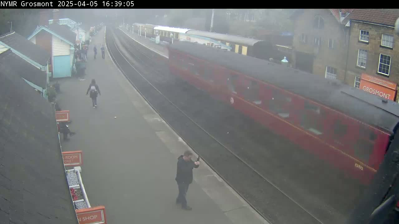 A red passenger train passes a railway platform on an overcast day, with several people waiting and walking nearby.