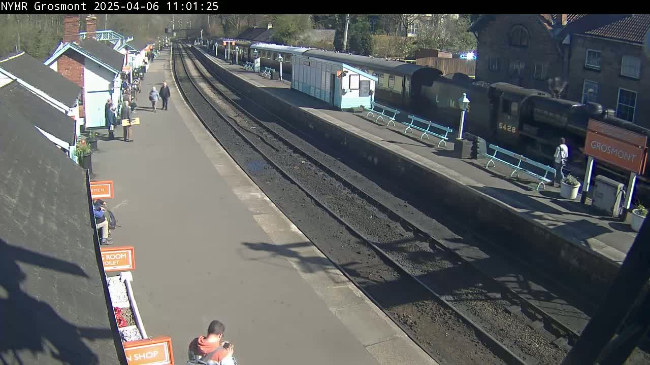 A sunny day at a train station shows a black steam engine and passenger cars at the platform with several people walking around.