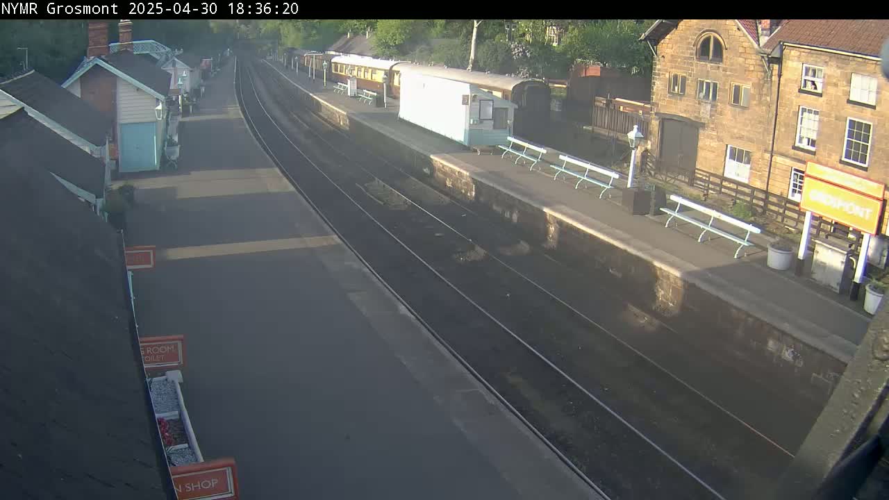 A train station platform with passenger train cars and several benches is visible on a partly sunny day.