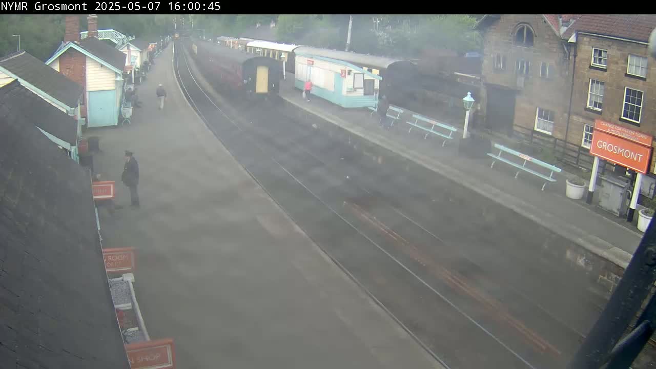A steam train sits at a station platform partially obscured by fog or steam, with several people standing around the area.