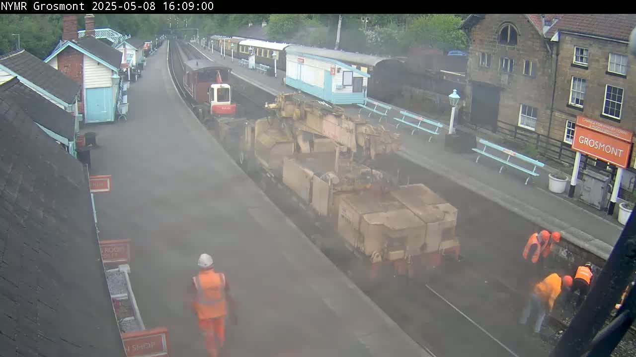 A railway platform with several workers in orange vests near a large piece of railway maintenance equipment, under a hazy sky.