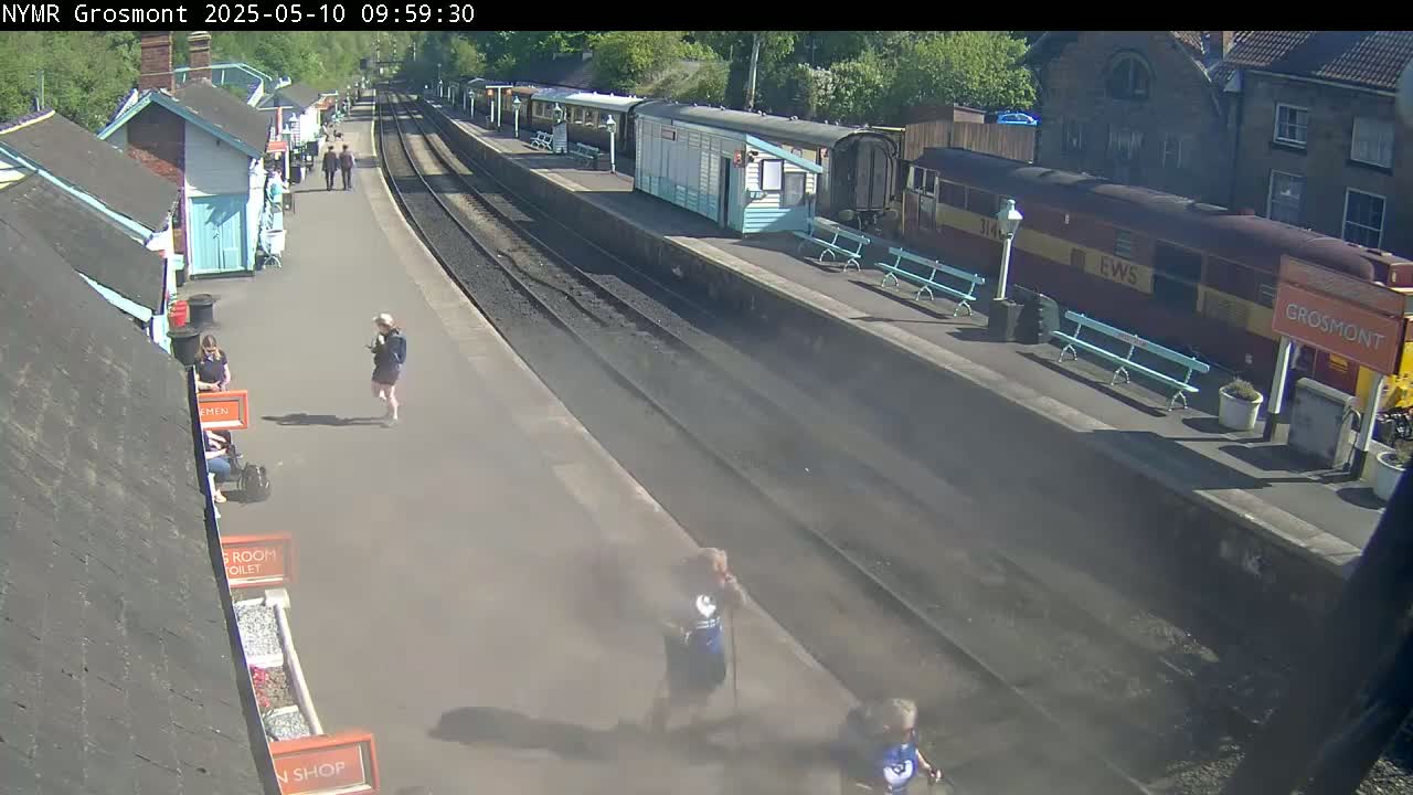 A sunny day at a train station shows several people walking near railway tracks and several trains parked at the platform.