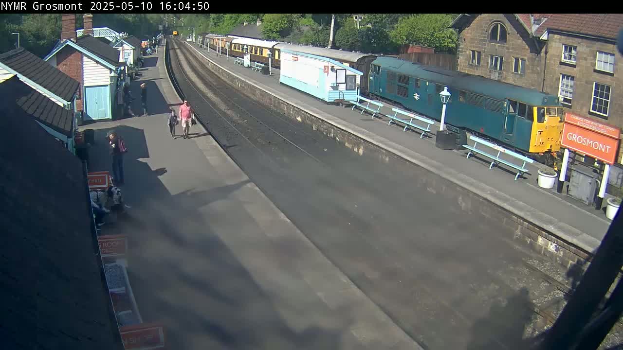 A sunny day at a train station shows several people walking on the platform near a teal and yellow diesel locomotive and several passenger cars.