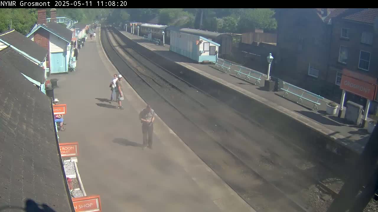 A sunny day at a train station shows several people walking on the platform near several small buildings and parked train cars.