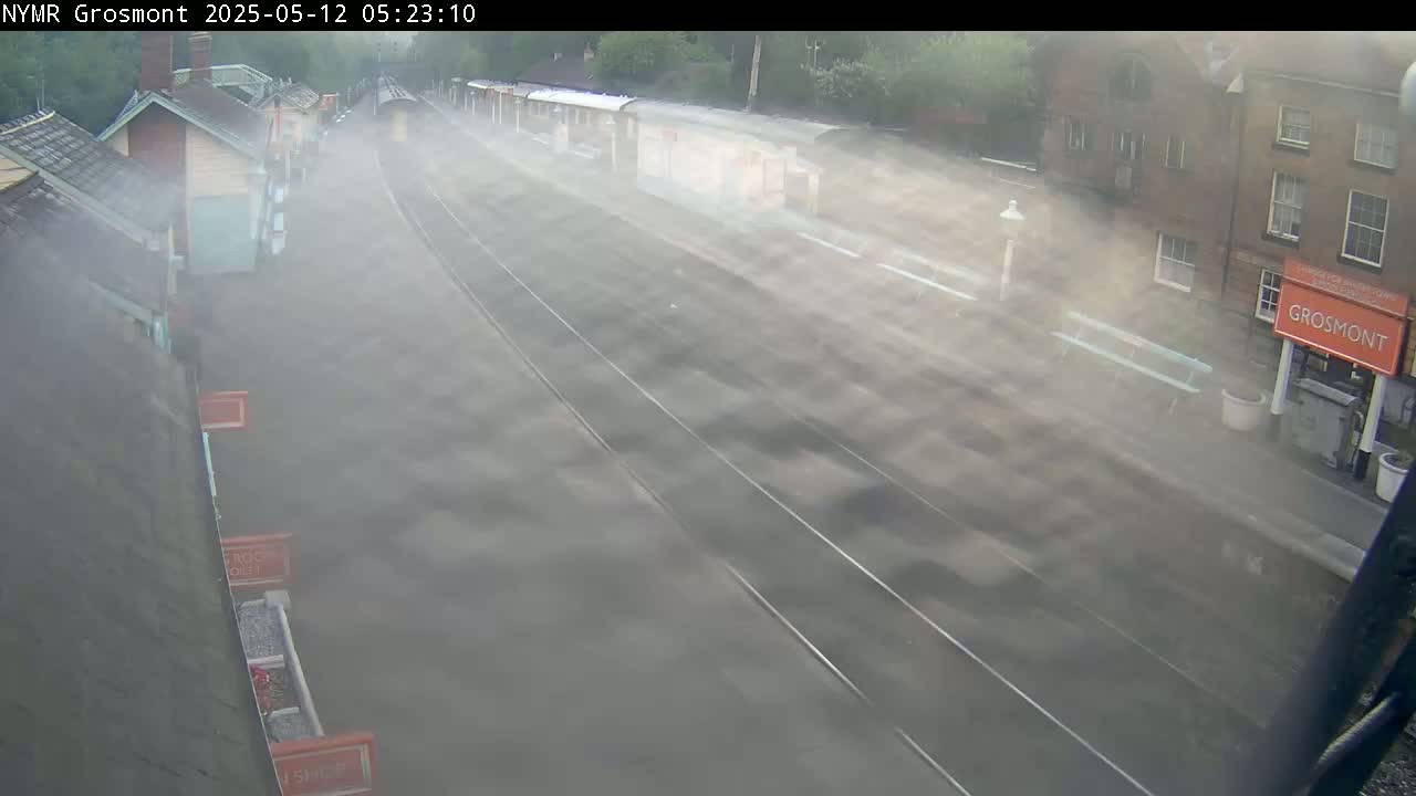 A train station platform is shrouded in a thick fog, with train cars partially visible in the background.