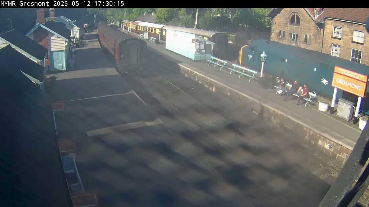 A steam train pulls into a station on a sunny day, with several people sitting on benches on the platform.