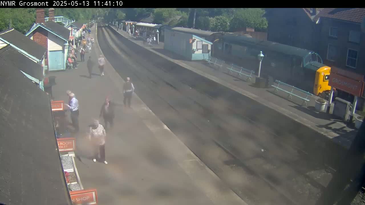 A train sits at a station platform on a sunny day, with several people walking on the platform and near some small buildings.