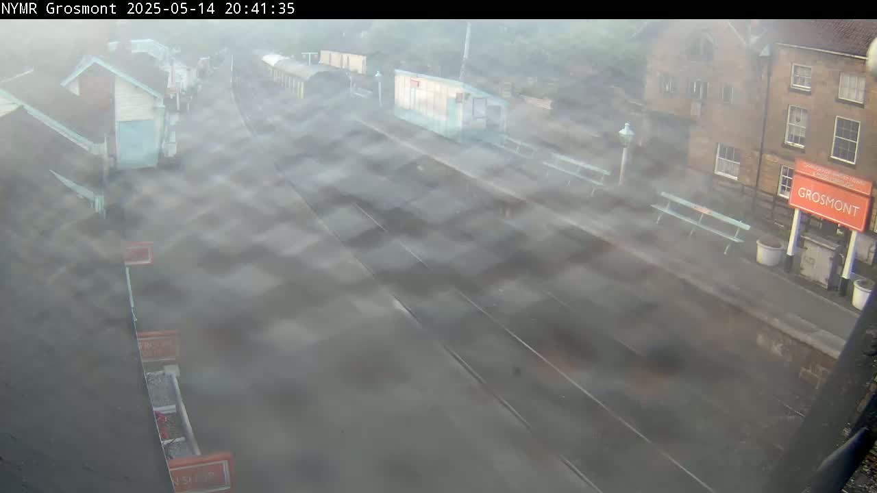 A hazy, possibly rainy, view of a train station with buildings, tracks, and benches.
