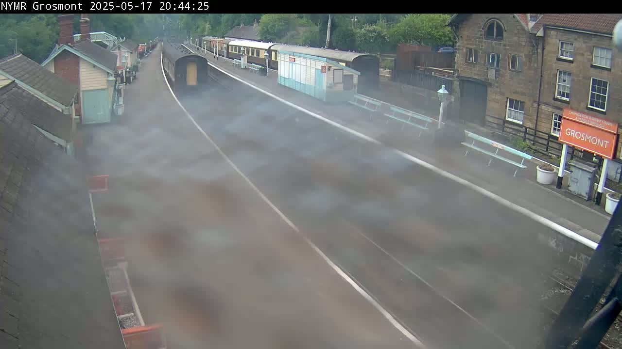 A train station platform is partially obscured by fog or steam, with trains visible in the background and station buildings nearby.