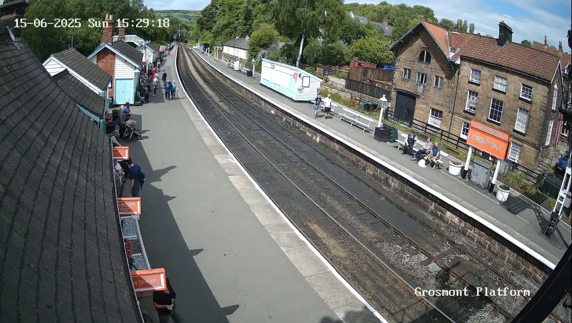 A high-angle view of a train station platform on a sunny day, showing people standing and sitting near small buildings and train tracks.