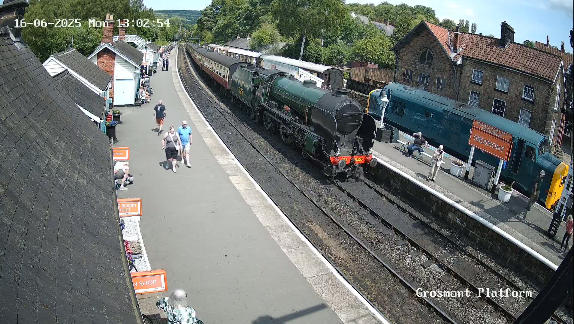 A steam train and a diesel locomotive are at a train station on a sunny day, with several people walking on the platform.