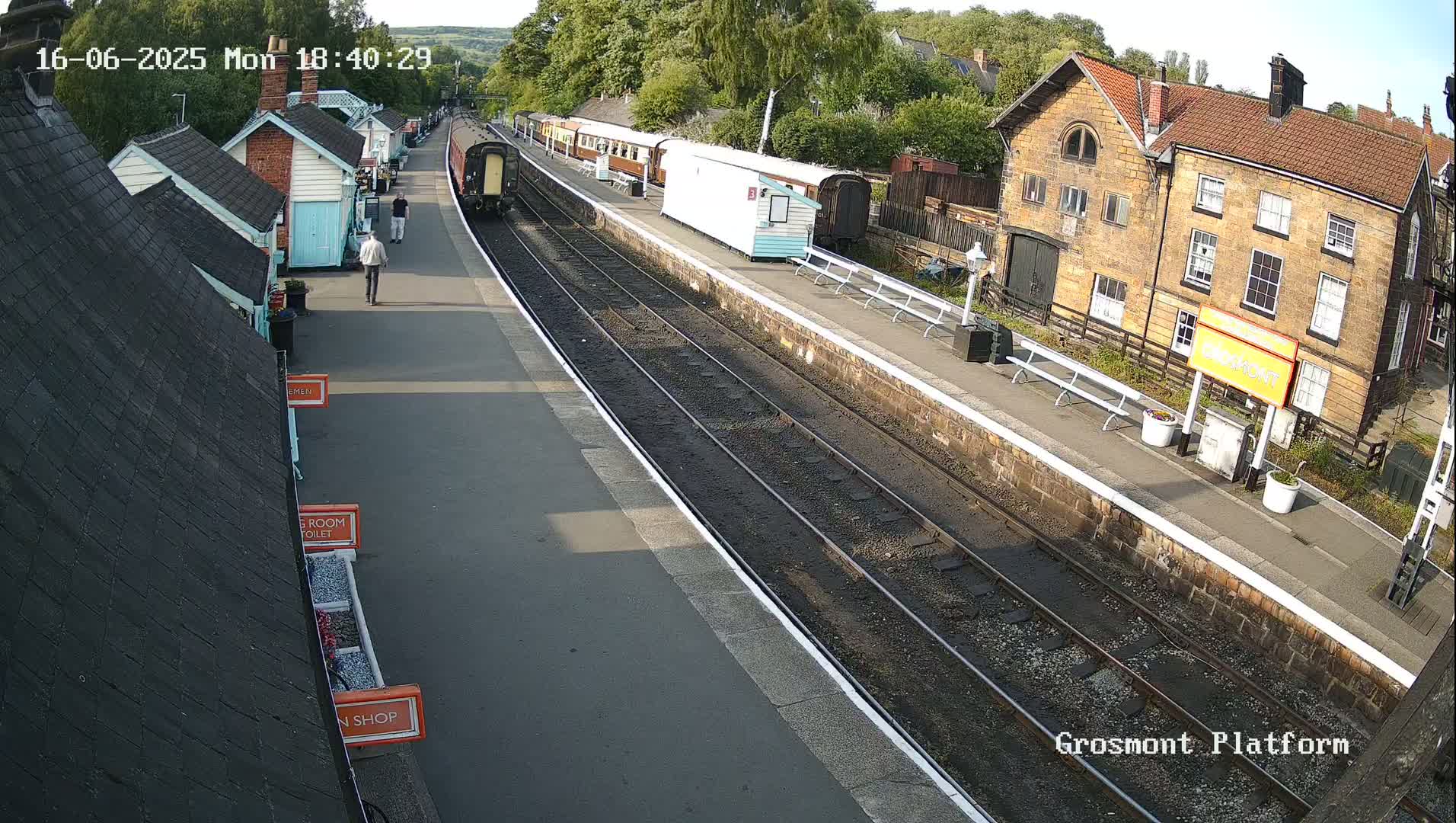 A train is arriving at a station on a sunny day, with several passenger cars and smaller buildings visible alongside the tracks.