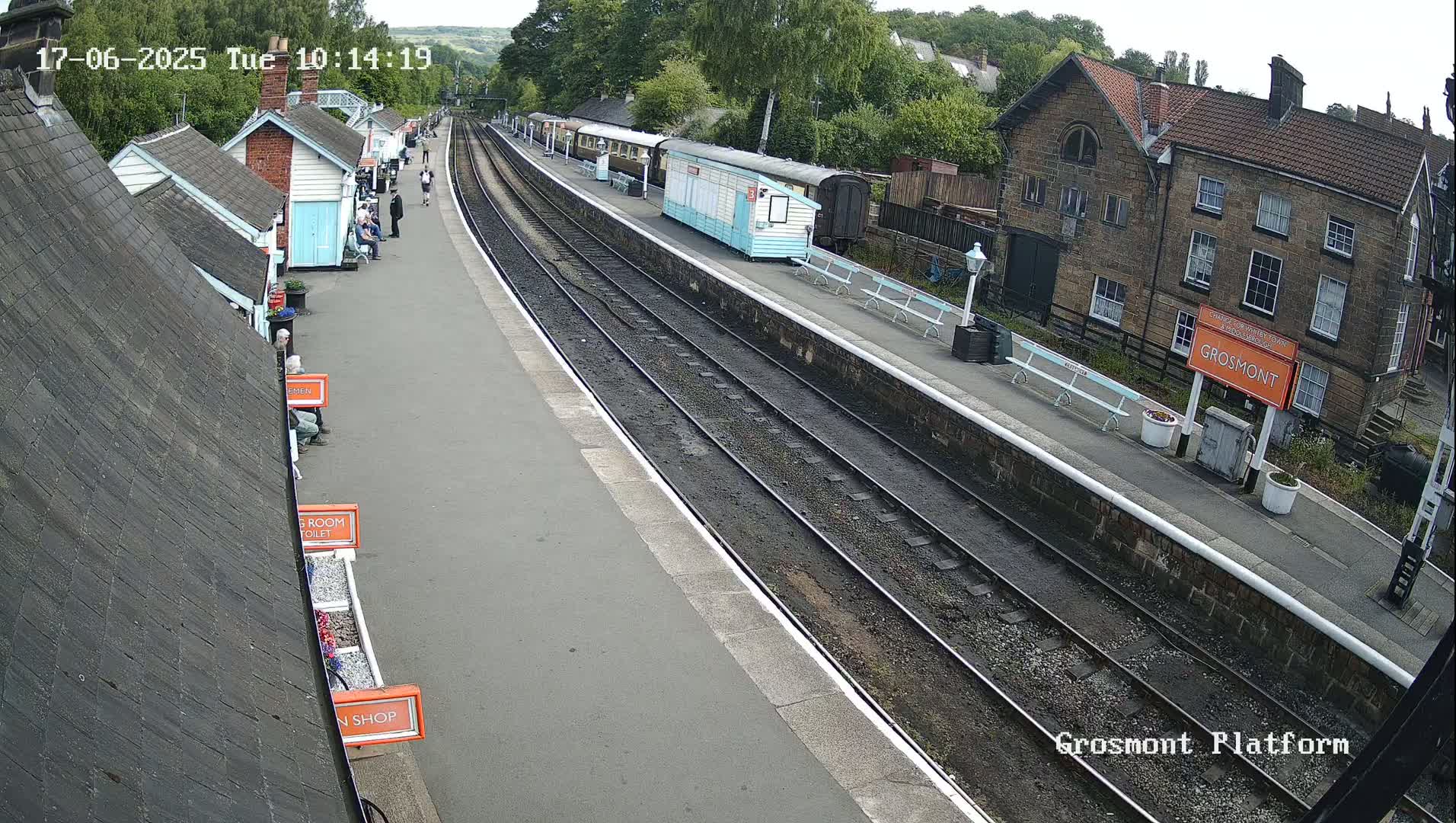 A train station platform with multiple train cars, several small buildings, and a few people on a partly sunny day.