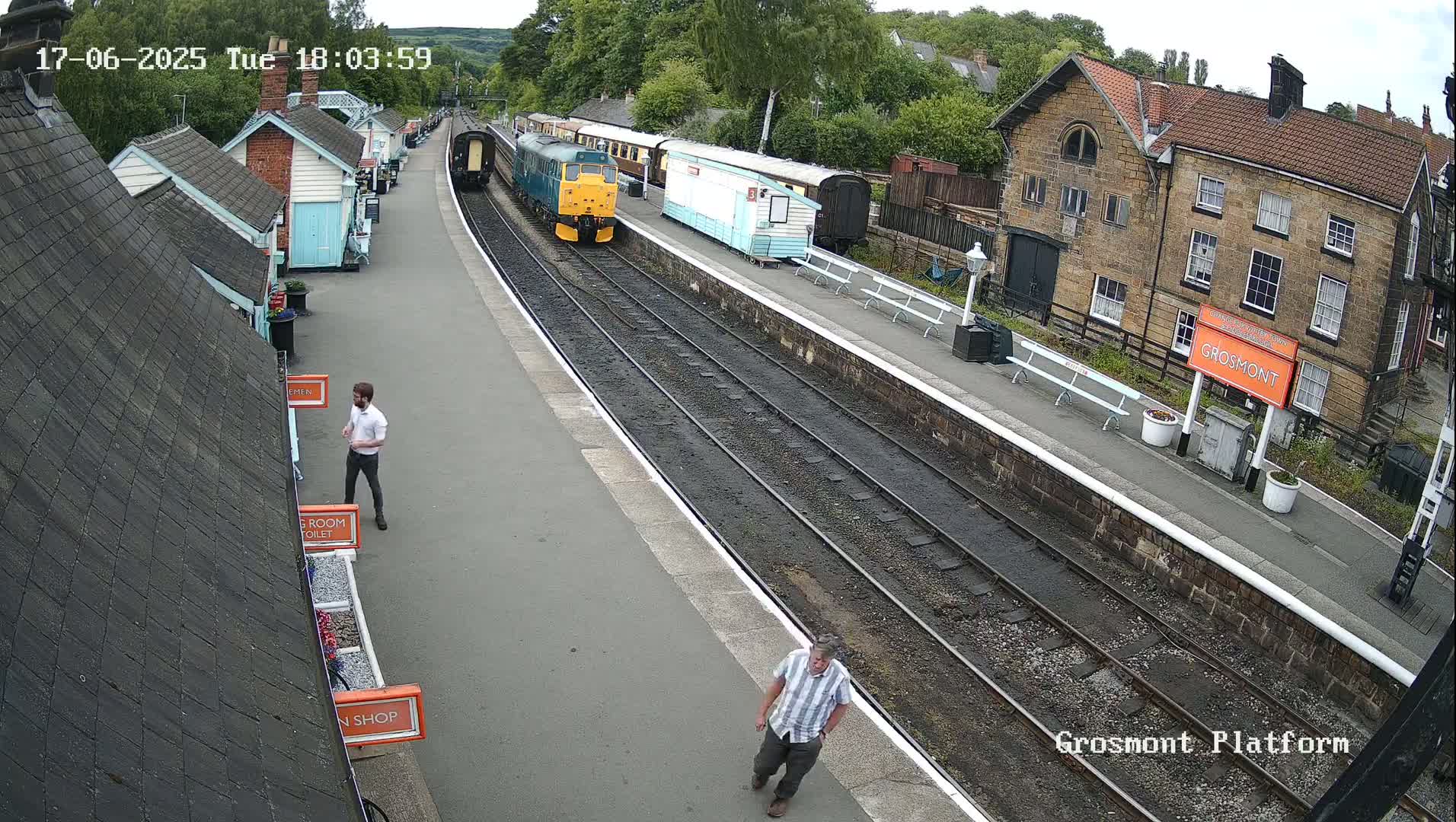 A train sits at a station platform on a partly sunny day; two people walk on the platform.