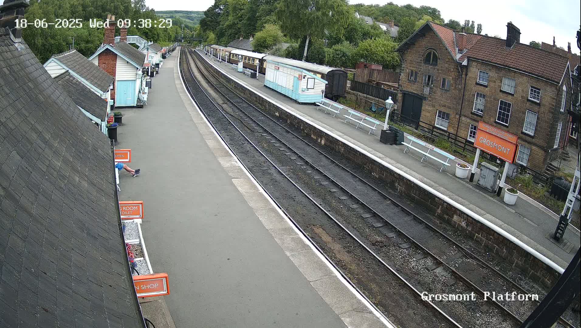 A railway platform with several passenger cars, small buildings, and a person sitting on a bench on a partly cloudy day.
