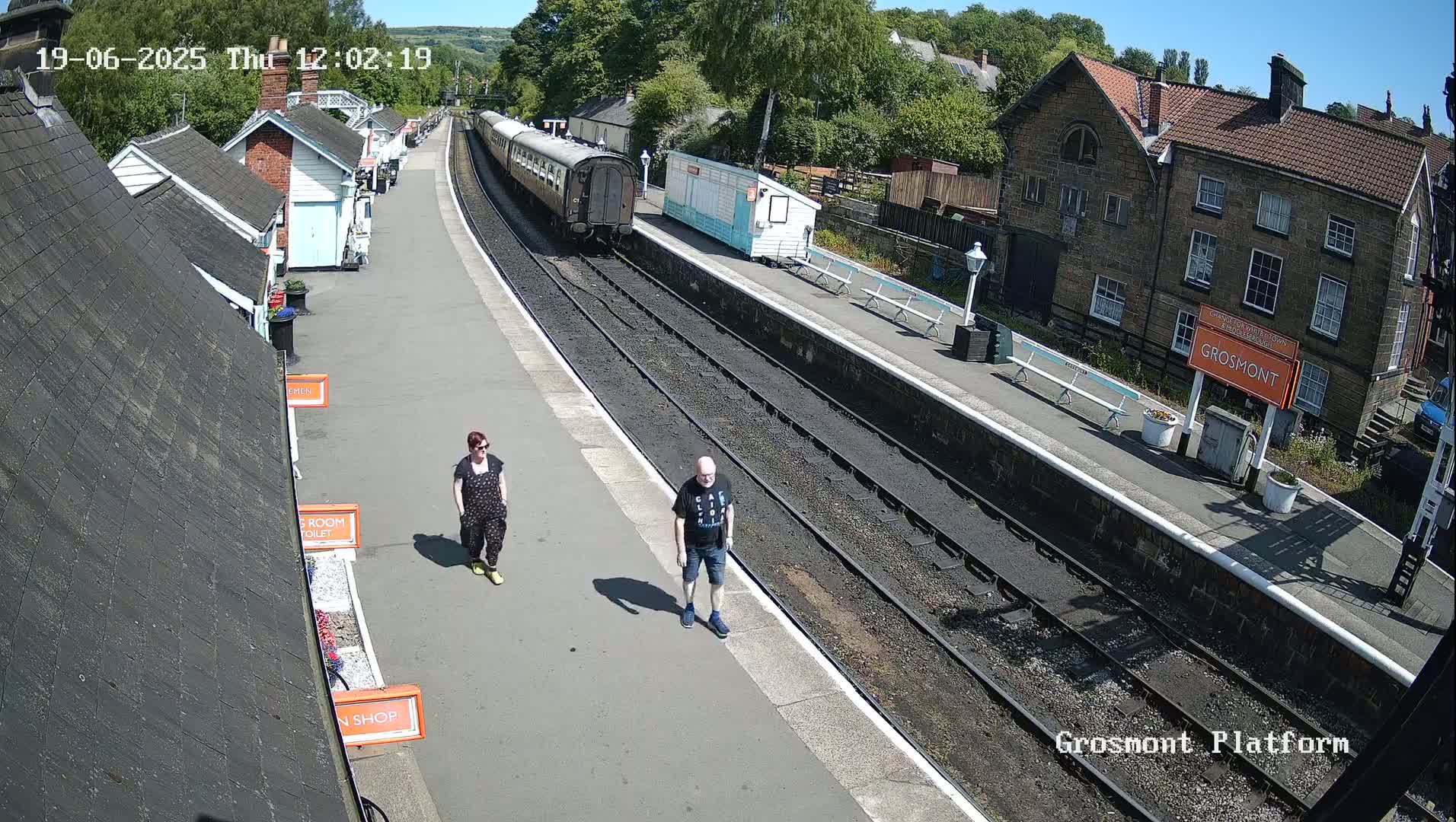 A man and a woman walk on a sunny train platform next to a train and several small buildings.
